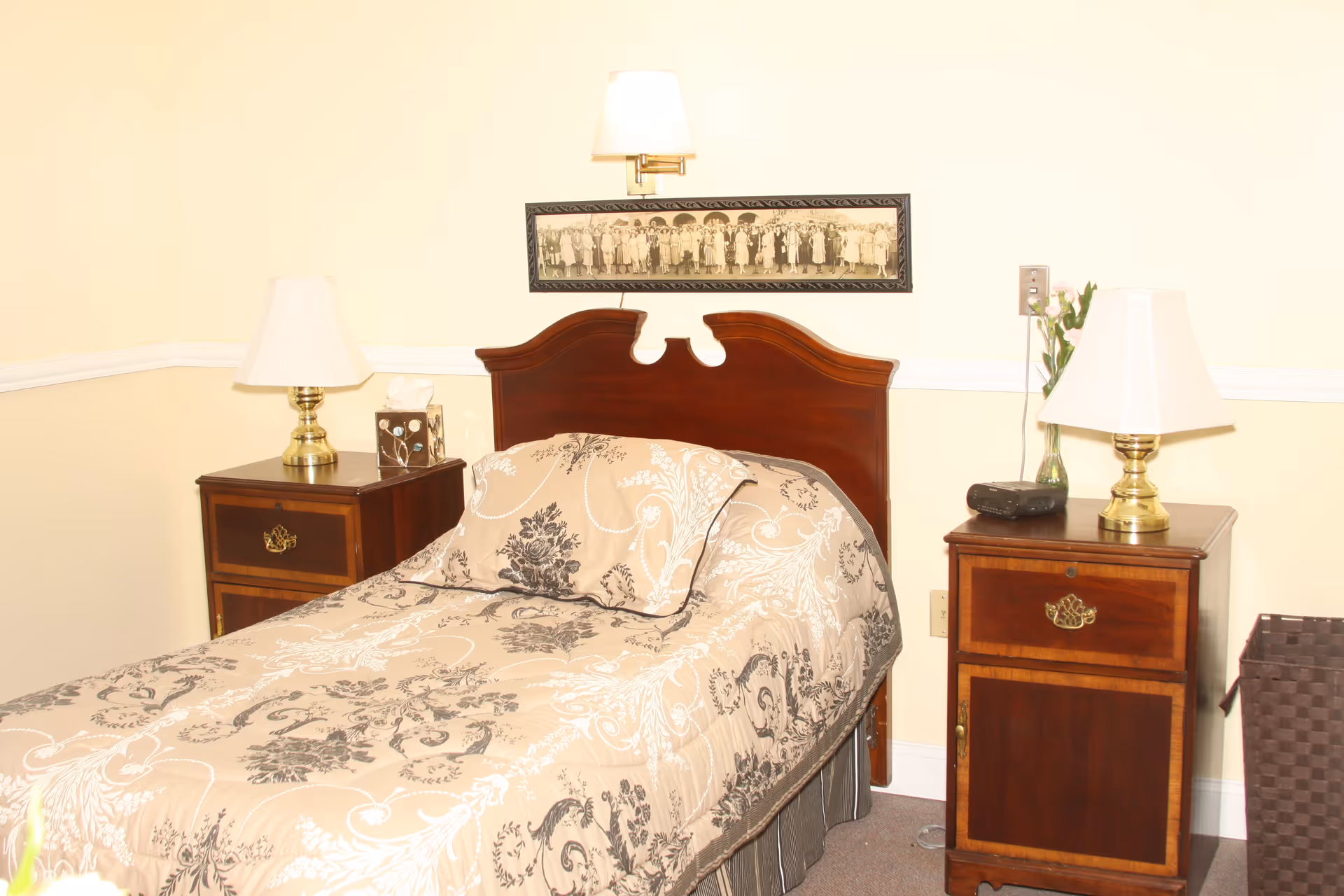 A bedroom with a single bed featuring a beige and black floral patterned comforter and pillow. The bed has a dark wooden headboard. On either side of the bed are matching wooden nightstands with brass lamps and various items including a tissue box and a small radio. Above the bed is a framed black and white photograph of a group of people. The walls are painted light yellow with white trim.