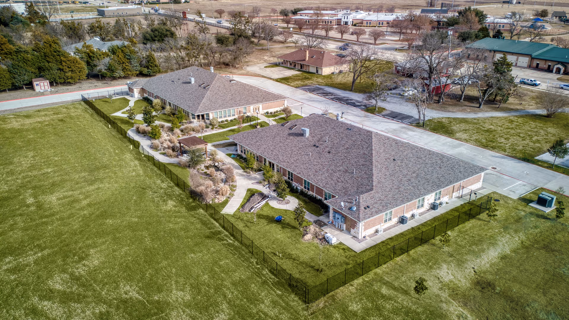 Aerial view of two single-story buildings with brown shingled roofs surrounded by green lawns and a fenced garden area. The buildings are part of a larger complex with paved roads and parking areas visible in the background. Trees and shrubs are scattered around the property.