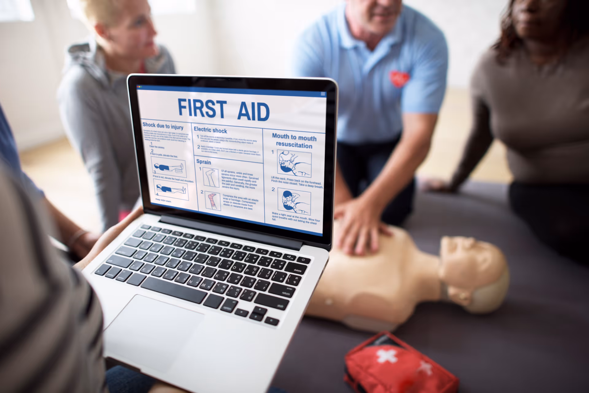 Laptop displays a first aid guide while trainees and a CPR mannequin practice nearby.