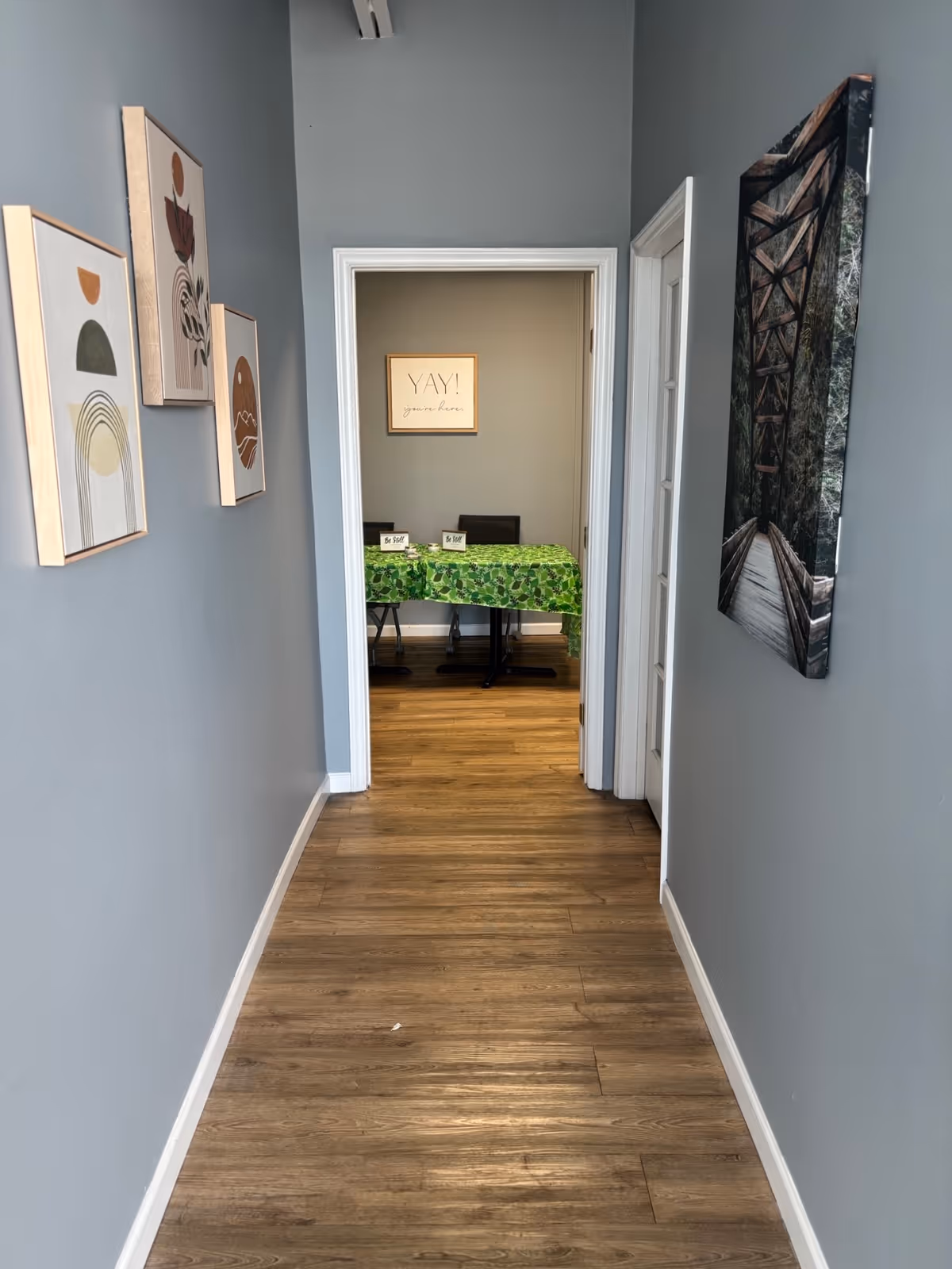 Narrow hallway with gray walls, wood floor, and art leading to a room with green table.