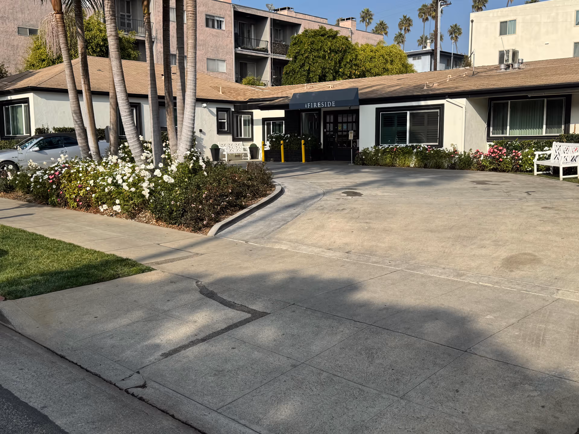 Exterior view of Fireside Health Care Center showing a single-story building with a beige roof and white walls. There is a driveway leading to the entrance with a black awning labeled 'FIRESIDE'. The area is landscaped with palm trees, bushes, and flowers. White benches are placed near the entrance.