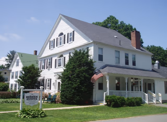 A large white multi-story house with black shutters and a gray roof, surrounded by green trees and bushes. An American flag is displayed on the front porch. A sign in front reads 'Berwick Estates Assisted Living Memory Care 79'. The sky is clear and blue.