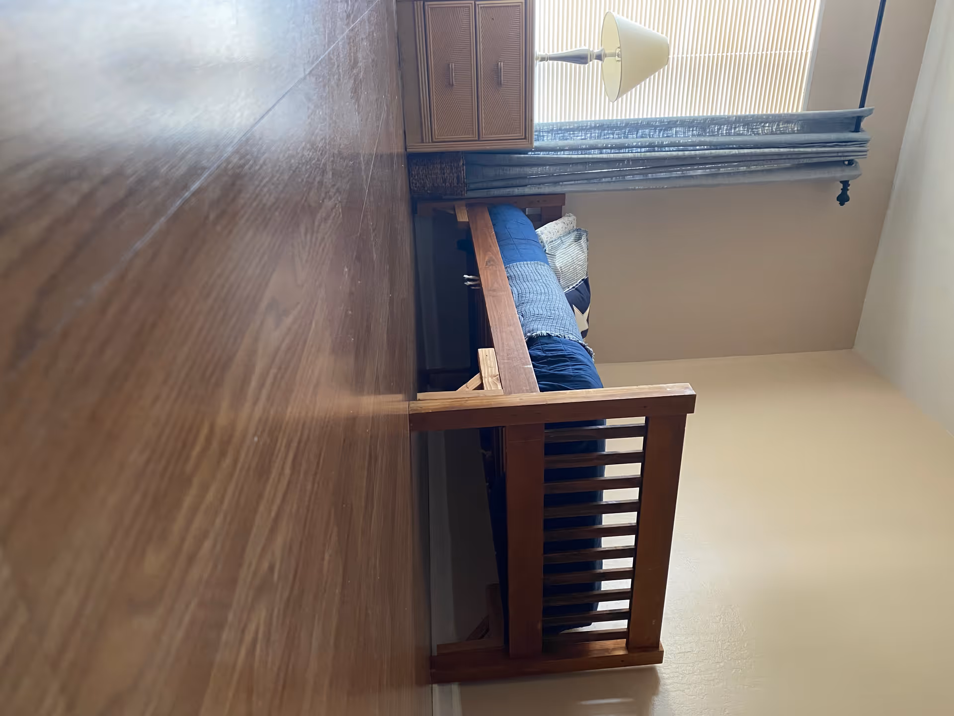 Wooden bed frame with blue bedding beside a window and lamp in a small bedroom.