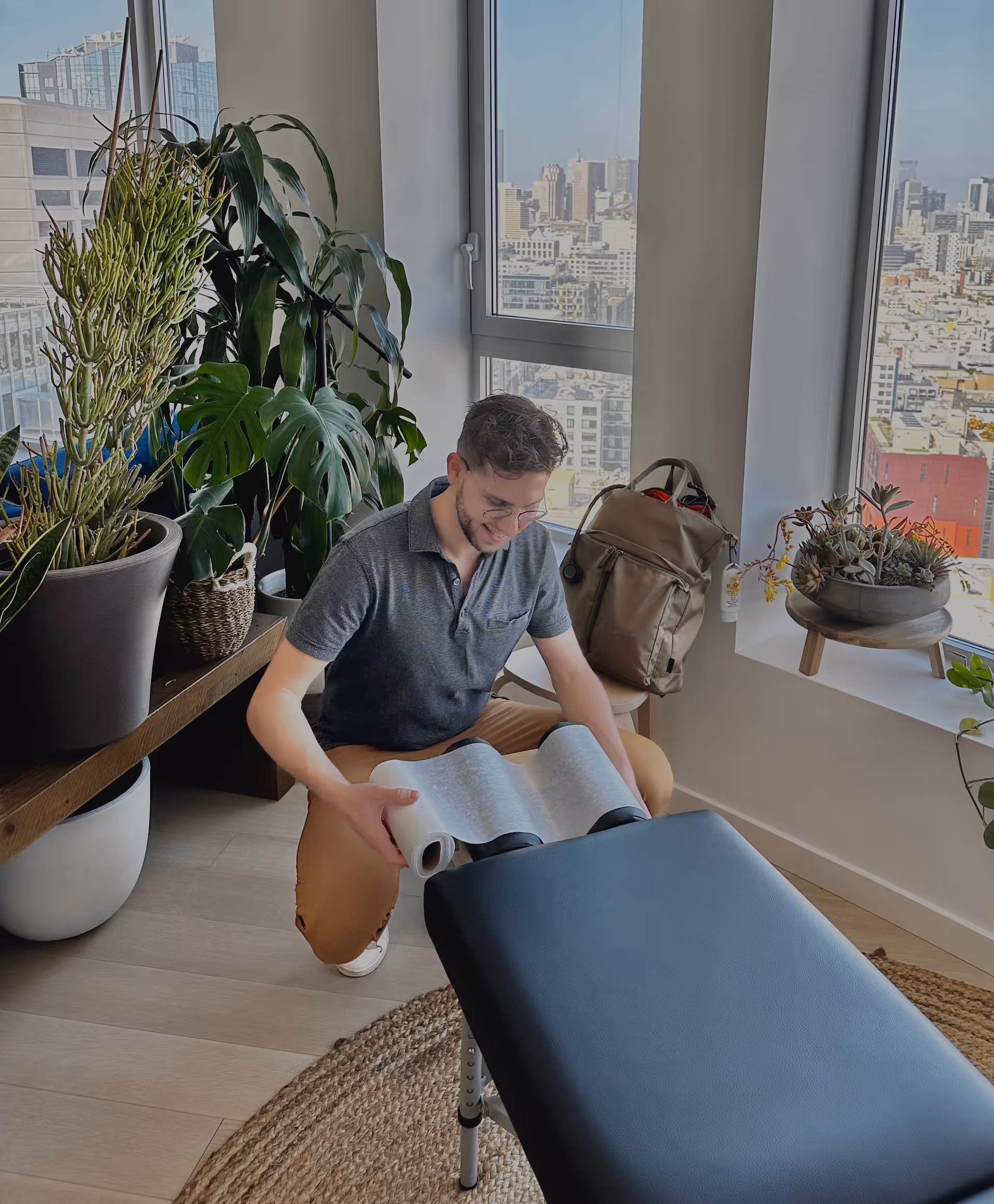 A man kneels beside a treatment table, unrolling paper in a bright, plant-filled clinic.