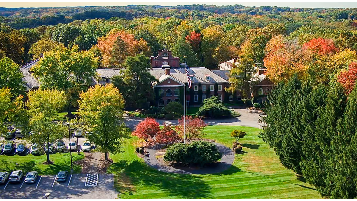 Aerial view of a senior living campus with a central brick building, flagpole, and colorful trees.