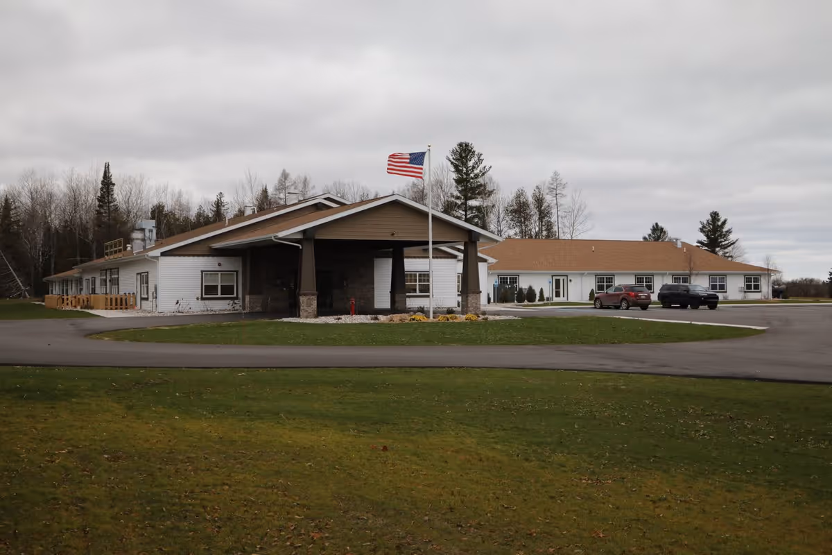 Exterior view of The Brook of Rogers City facility showing a single-story building with a covered entrance supported by pillars, an American flag on a flagpole in front, a paved driveway, two parked vehicles, and a grassy area in the foreground under a cloudy sky.