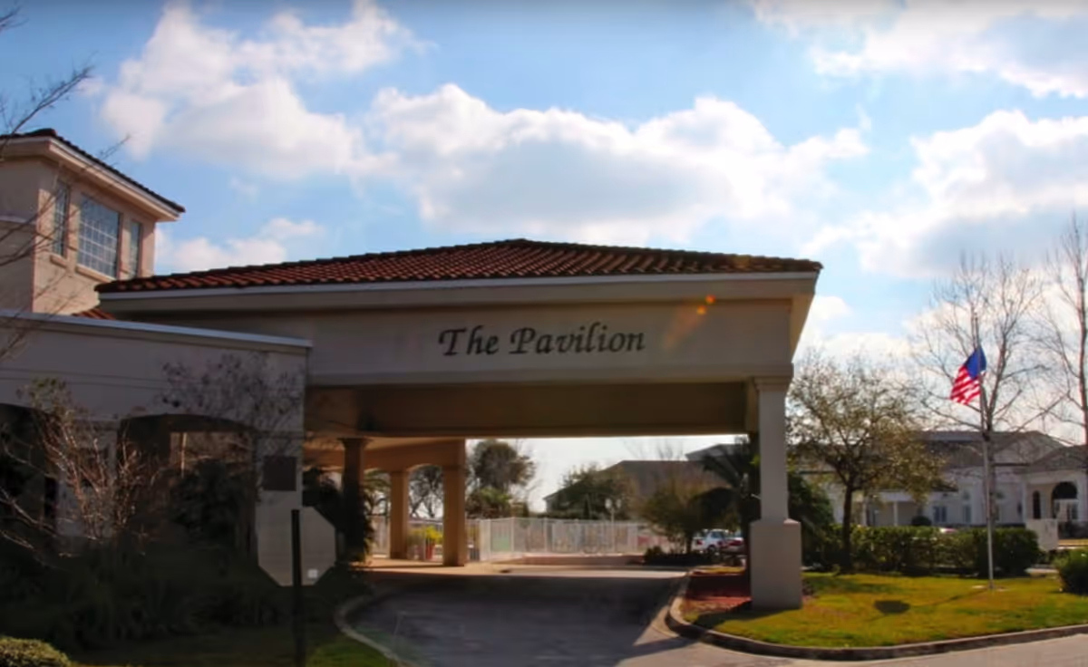 Drive-through entrance canopy labeled "The Pavilion" at the front of a building with an American flag and partly cloudy sky.