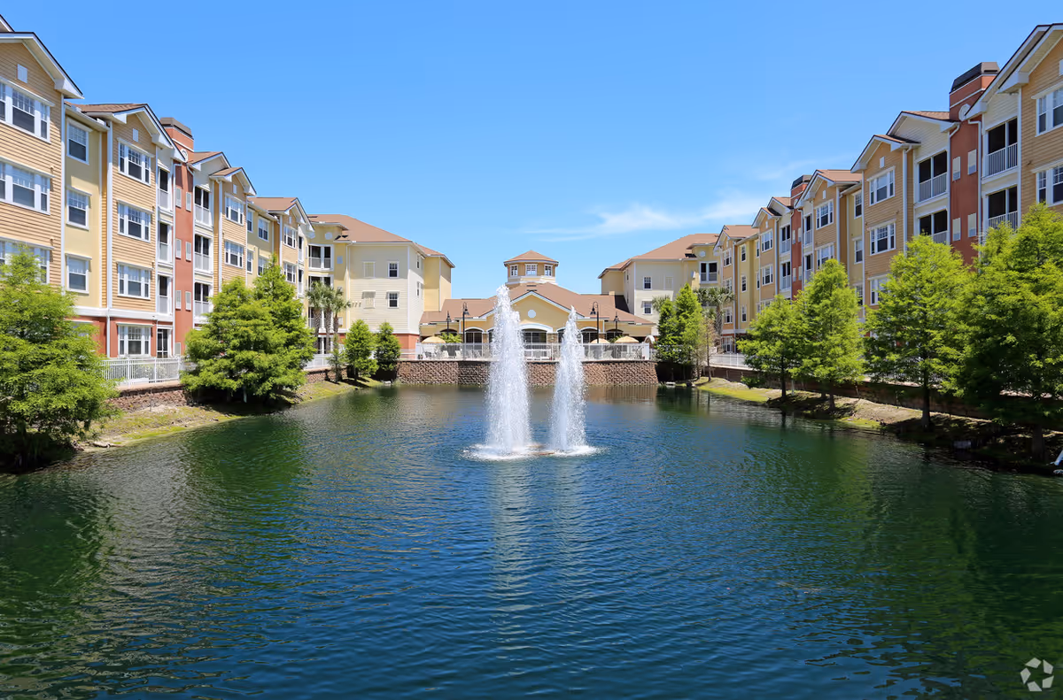 Colorful multi-story senior living buildings surround a central pond with a fountain.