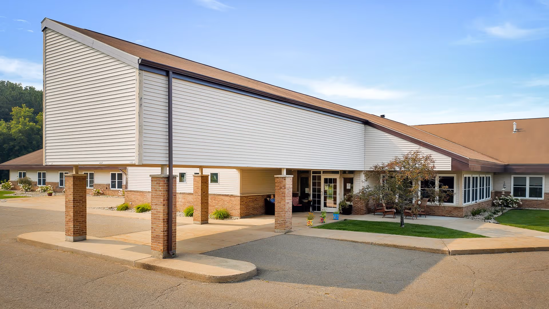 Exterior view of Wellington Place At Rib Mountain showing the building entrance with a covered drop-off area supported by brick pillars, surrounded by a paved driveway and landscaped greenery under a clear blue sky.