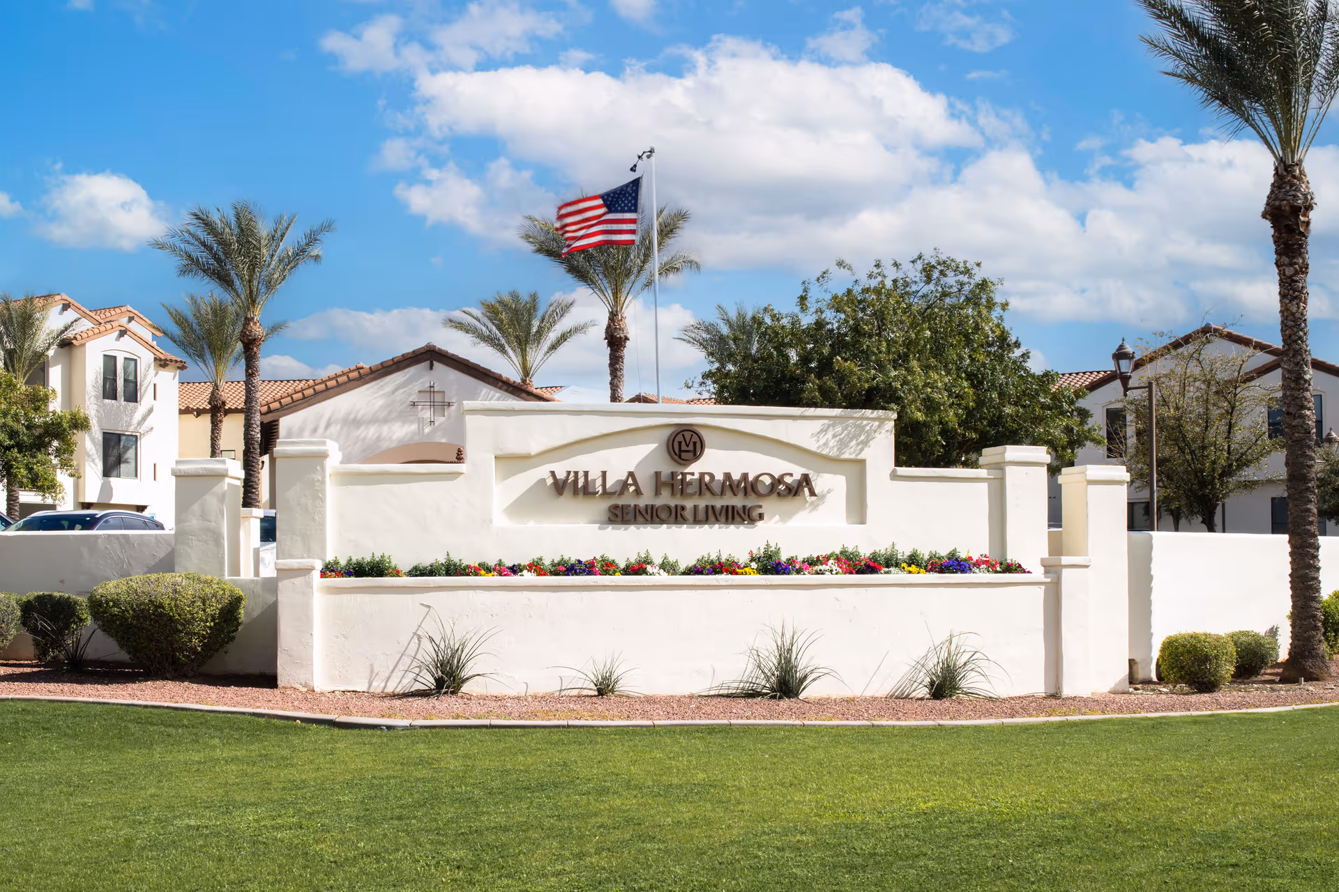 Exterior view of Villa Hermosa Senior Living facility entrance sign with landscaping, palm trees, and an American flag against a blue sky with clouds.