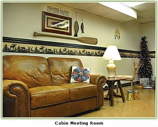 A cozy meeting room with a brown leather couch, a wooden side table with a lamp, a chair, and a small decorated Christmas tree. The wall has rustic decor including a wooden paddle, framed pictures, and a welcome sign. The room has a warm, cabin-like atmosphere.