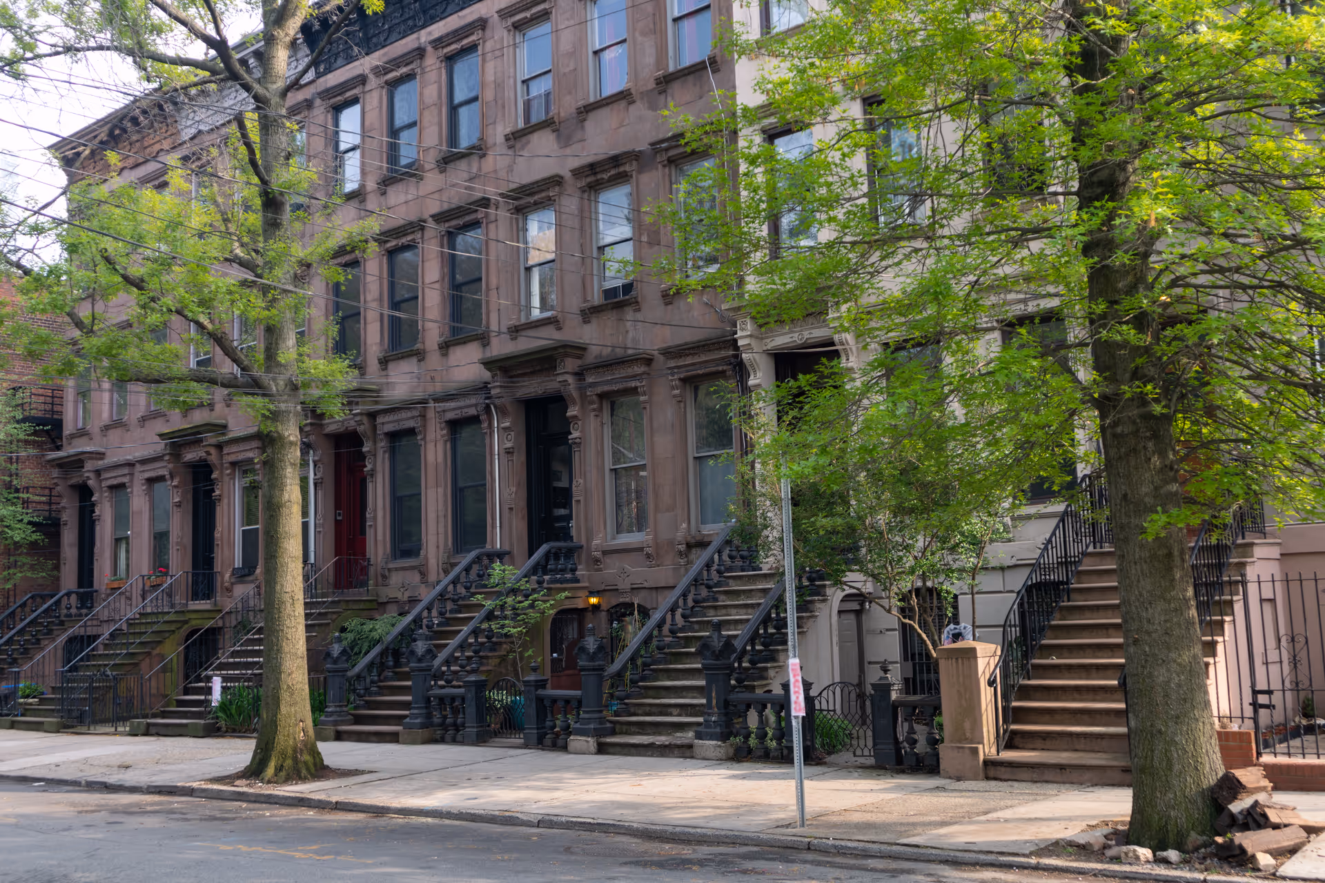 Row of brownstone townhouses with stairs, railings, and leafy trees along a city street.