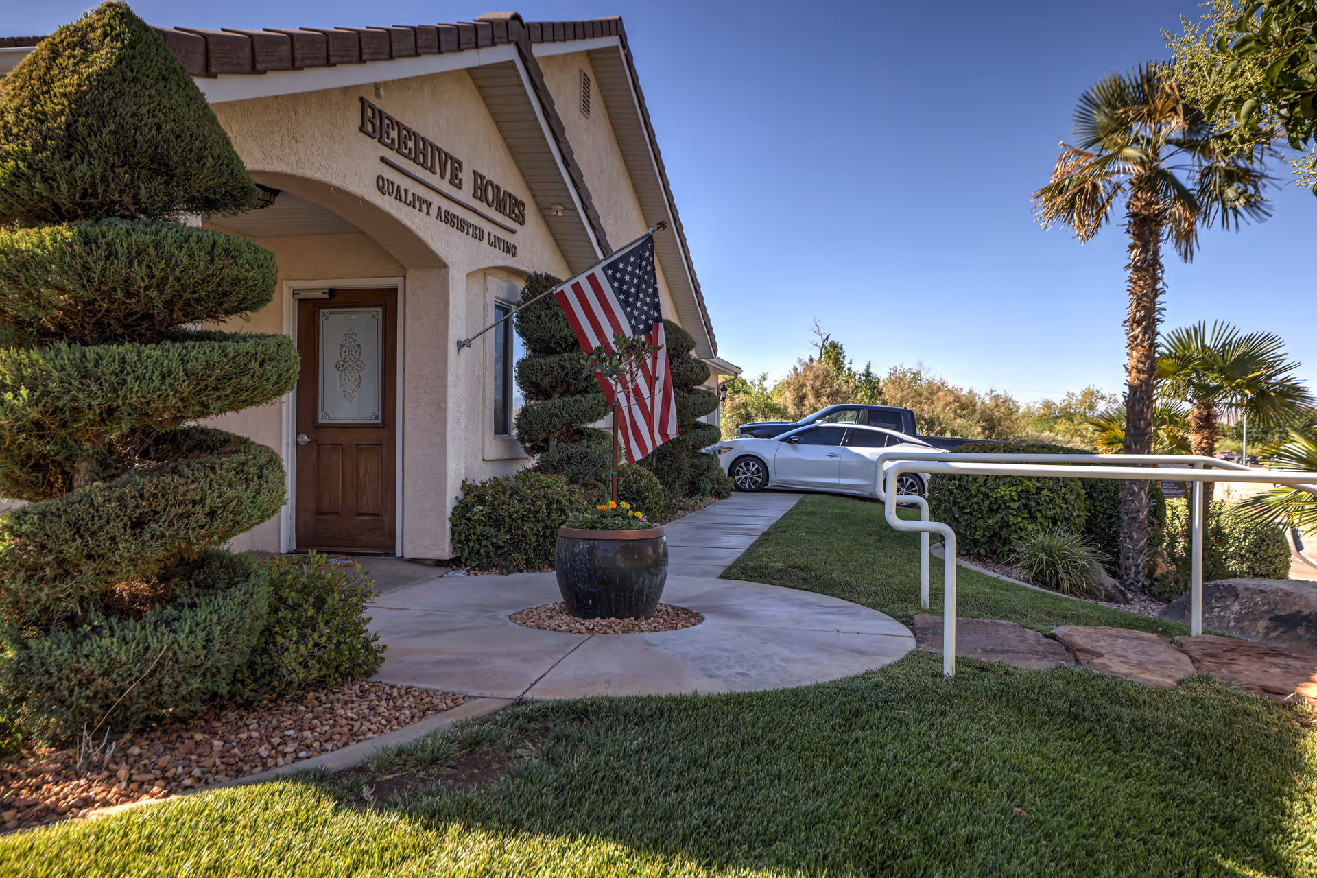 Front entrance of a Beehive Homes memory care building with an American flag, sculpted shrubs, a potted plant, and an accessibility ramp.
