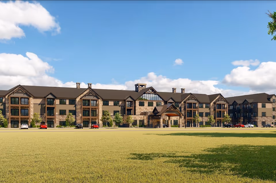 Exterior view of a large, multi-story senior living facility building with stone and wood accents, multiple windows, and a covered entrance. Several cars are parked in front, and there is a large grassy area in the foreground under a partly cloudy sky.