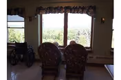 A sunlit living room with two upholstered chairs by large windows and a wheelchair.