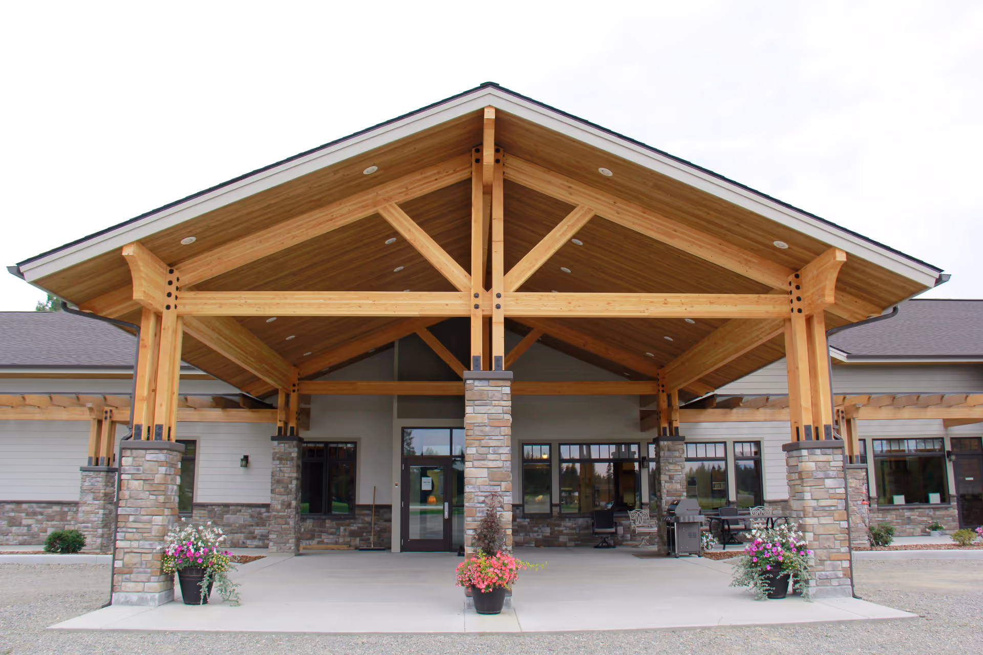 Front entrance of an assisted living building with a large wooden covered porte-cochère supported by stone columns and potted flowers.