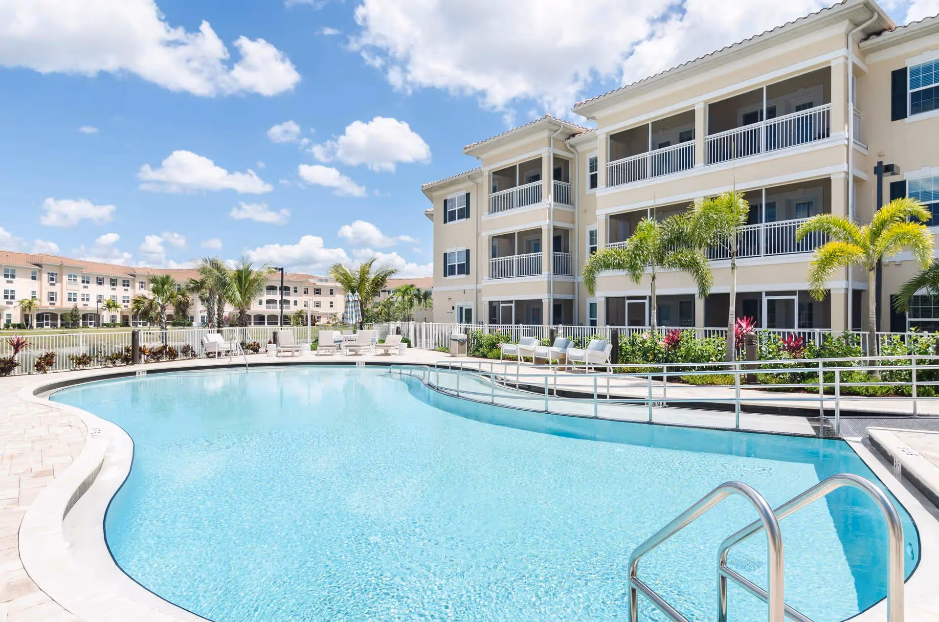Sunlit outdoor pool area with lounge chairs beside a beige senior living building.