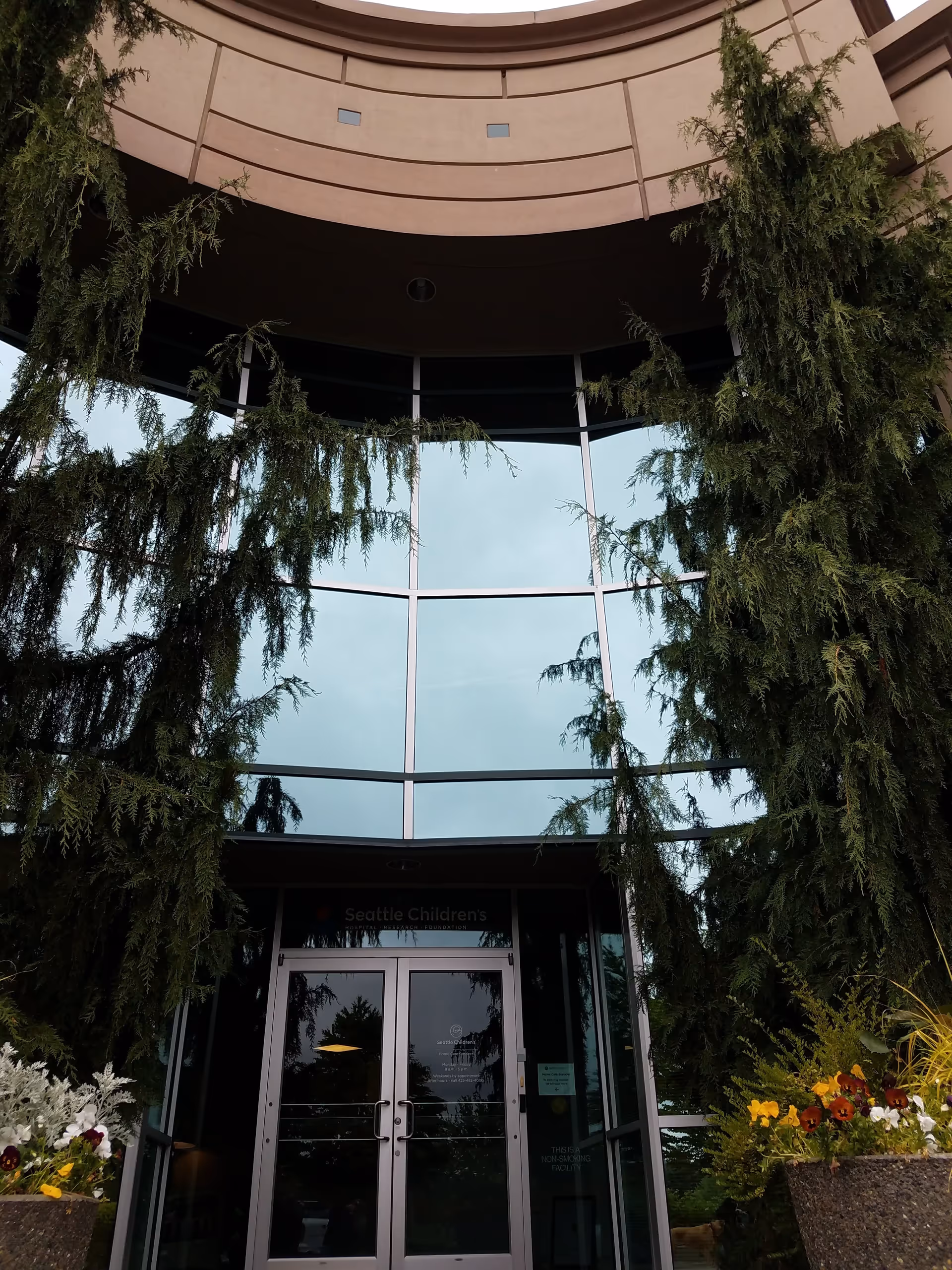 Curved glass entrance to Seattle Children's Home Care Services, framed by tall evergreen trees.
