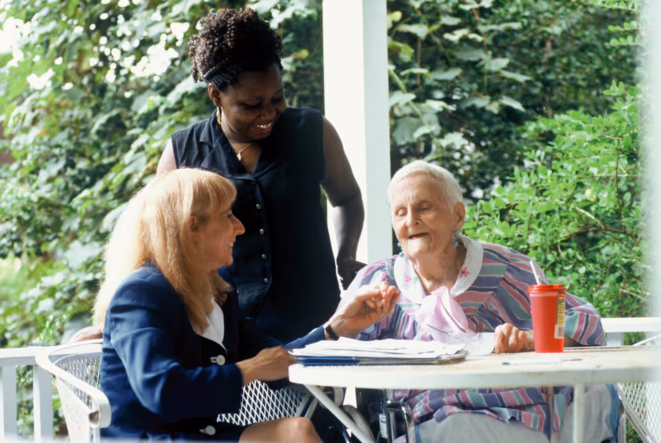 Three people gather at an outdoor porch table, smiling and chatting.