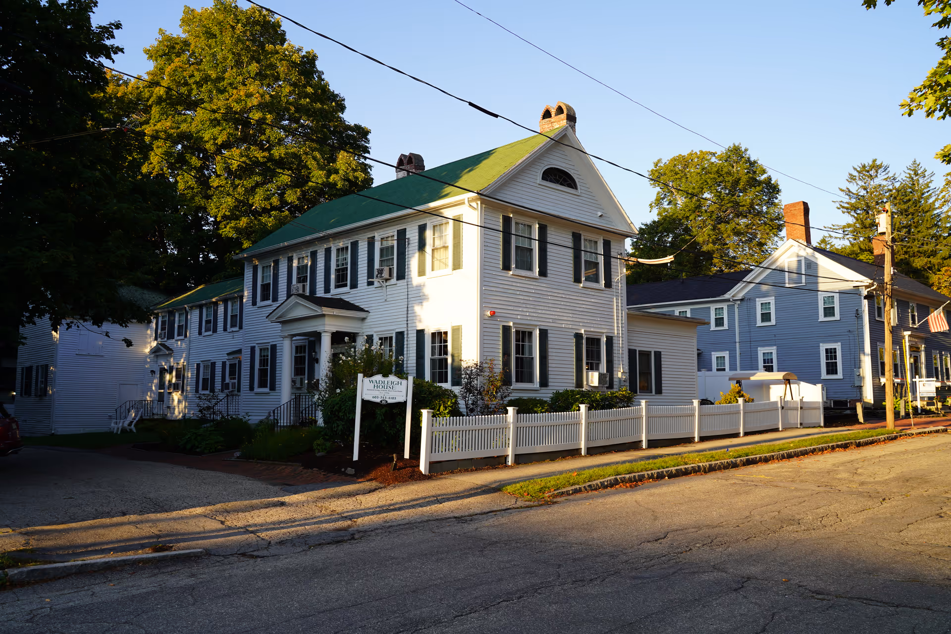 White two-story building with green roof along a tree-lined street.