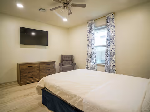 A tidy bedroom featuring a large bed, dresser, wall TV, chair, and patterned curtains.