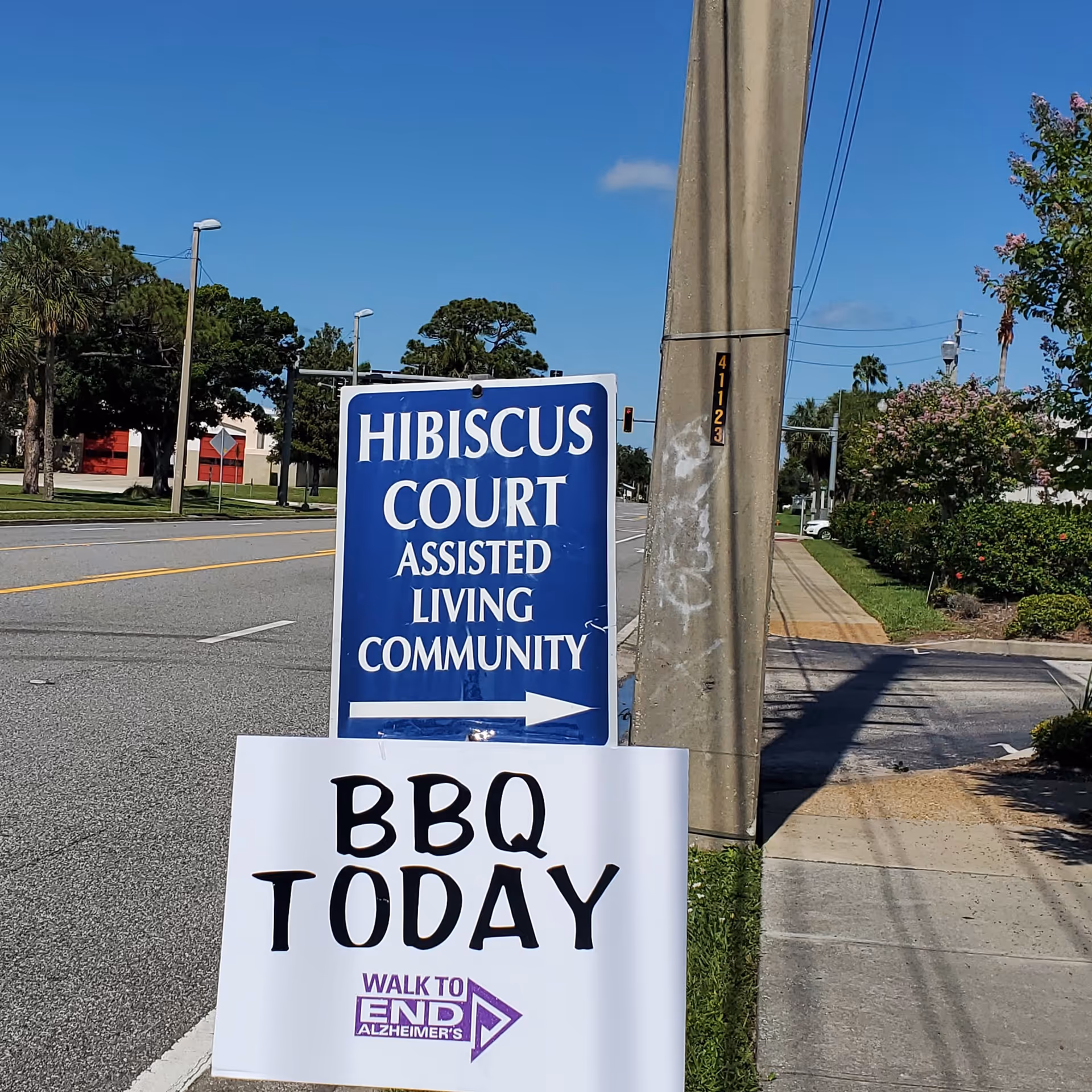 Blue Hibiscus Court Assisted Living Community sign with BBQ Today sign on a street corner.