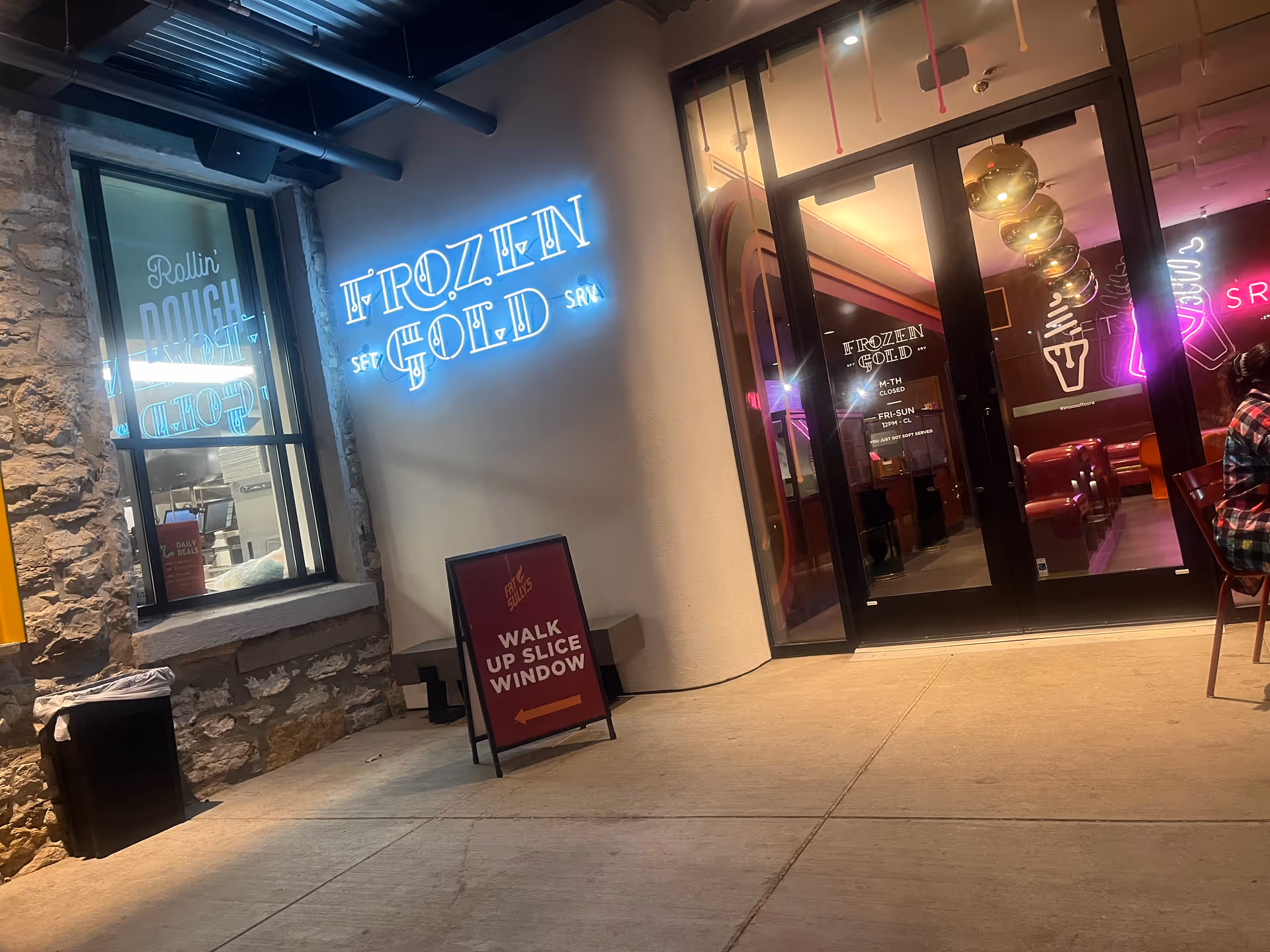 Exterior storefront with neon signs and glass doors, stone wall on left and sidewalk.