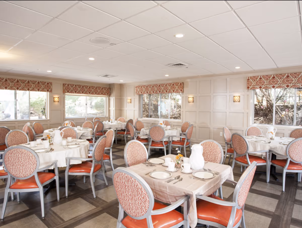 A bright dining room with multiple round tables covered with white tablecloths, each set with plates, cups, and silverware. The chairs have red cushioned seats and patterned backs. Large windows with patterned valances let in natural light, and the walls are paneled in a light color.