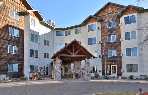 Front exterior view of a multi-story senior living facility with a combination of wood and light-colored siding. The building features multiple windows, a covered entrance supported by stone pillars, and an American flag near the entrance.