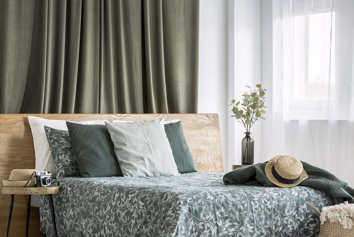 A neatly made bed with a wooden headboard, green and white patterned bedding, and multiple pillows in shades of green and white. A small side table next to the bed holds a straw hat and a vintage camera. A vase with green foliage is placed on a nightstand near a window with sheer white curtains, and a green blanket with a straw hat rests on the bed.