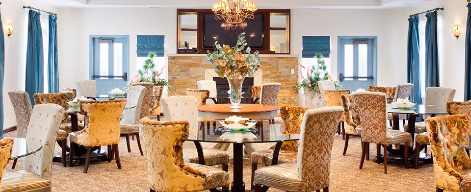 A bright and elegant dining room with multiple round glass tables surrounded by upholstered chairs in various floral and patterned fabrics. The room features large windows with blue curtains, a stone fireplace with a large mirror above it, and a chandelier hanging from the ceiling. Decorative plants and floral arrangements are placed around the room.