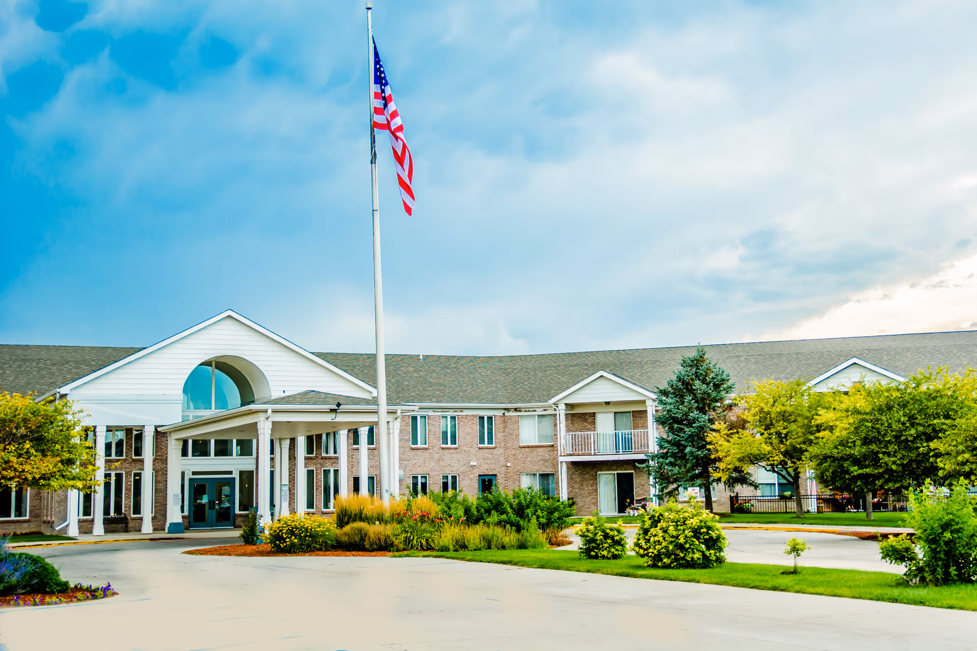 Front exterior view of a two-story retirement community building with a covered entrance, an American flag on a tall flagpole, and well-maintained landscaping including trees and shrubs under a partly cloudy sky.