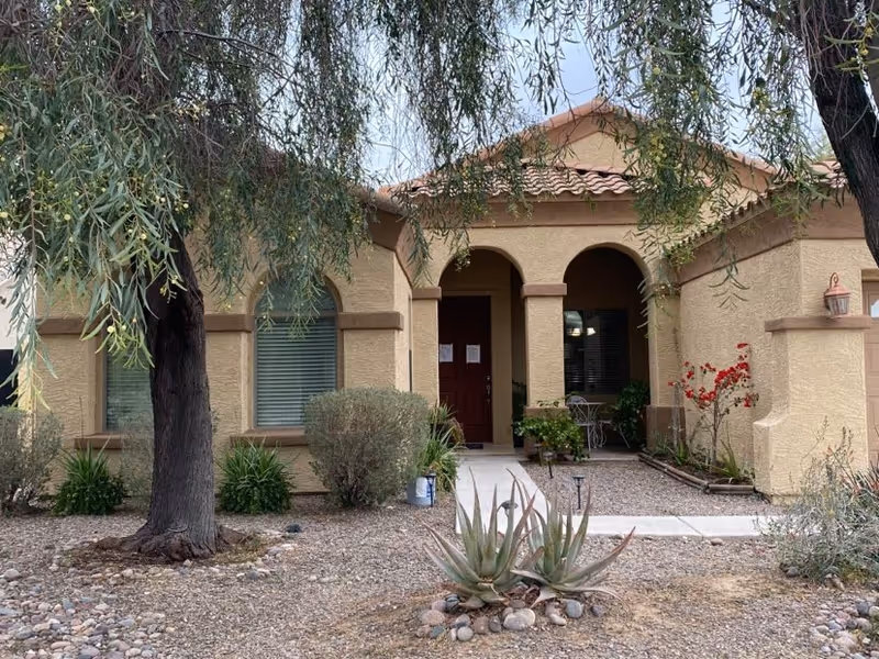 Front entrance of a stucco single-story building with an arched porch, a red door, potted plants, and desert landscaping with trees and succulents.