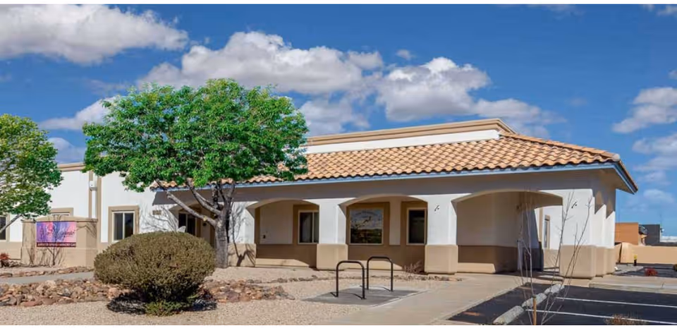Exterior view of a single-story building with a tiled roof and arched entryways, surrounded by desert landscaping including trees, bushes, and rocks under a partly cloudy blue sky.
