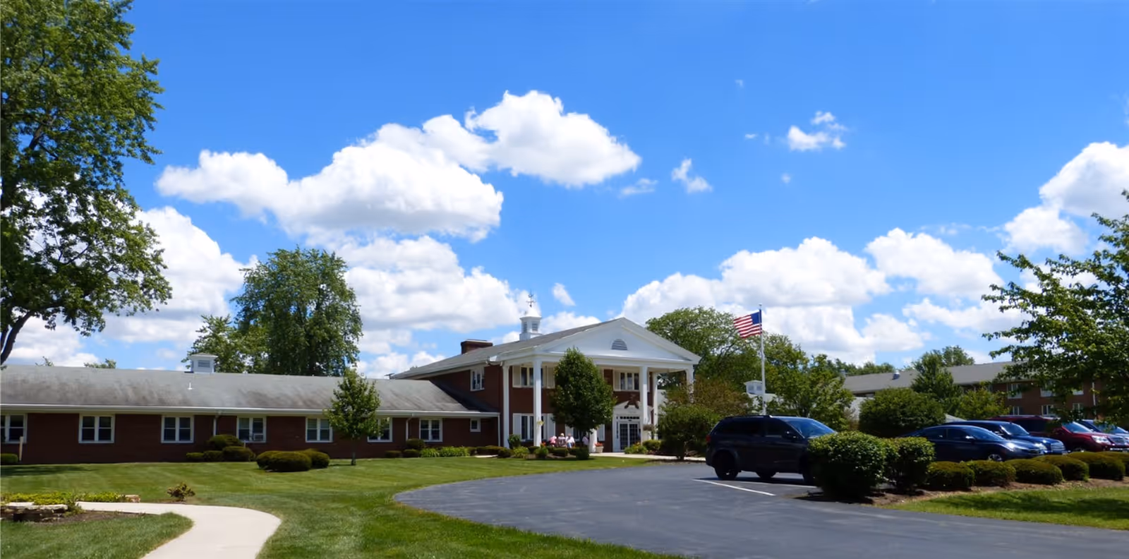 Front exterior of a single-story brick convalescent home with a columned entrance, American flag, parked cars and a manicured lawn under a blue sky.