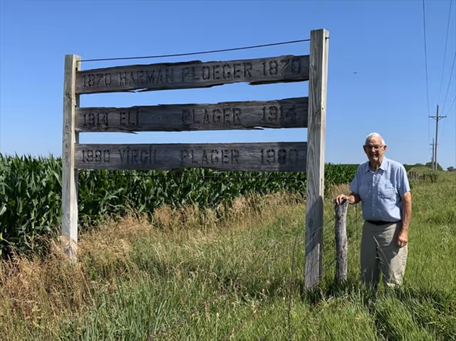 Elderly man stands beside a weathered wooden sign in a rural field.