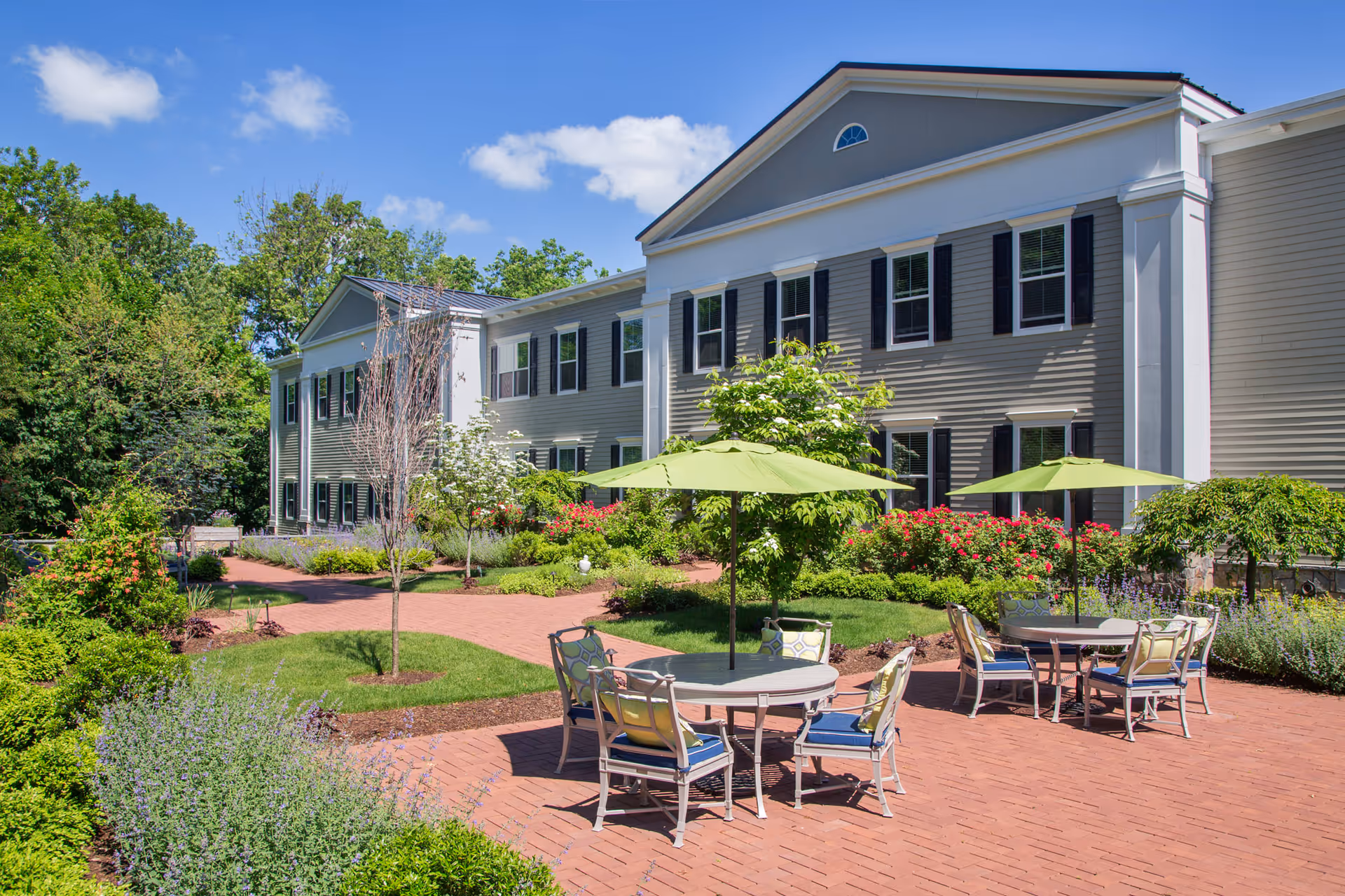 Outdoor patio area at a senior living facility with round tables, chairs, and green umbrellas on a brick-paved surface surrounded by landscaped gardens and a two-story building with white siding and black shutters in the background under a blue sky.