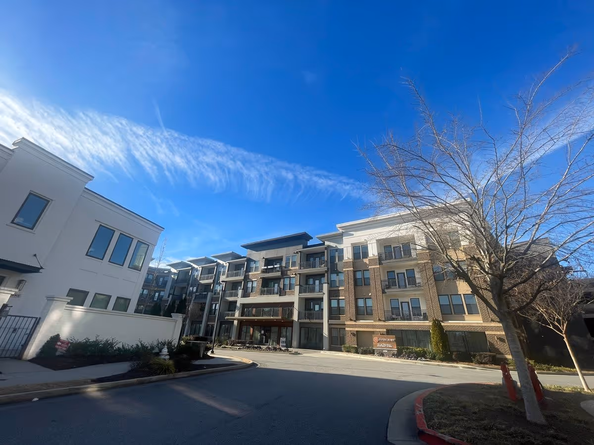 Front exterior of a multi-story apartment building with balconies, a driveway entrance, and a leafless tree under a bright blue sky.