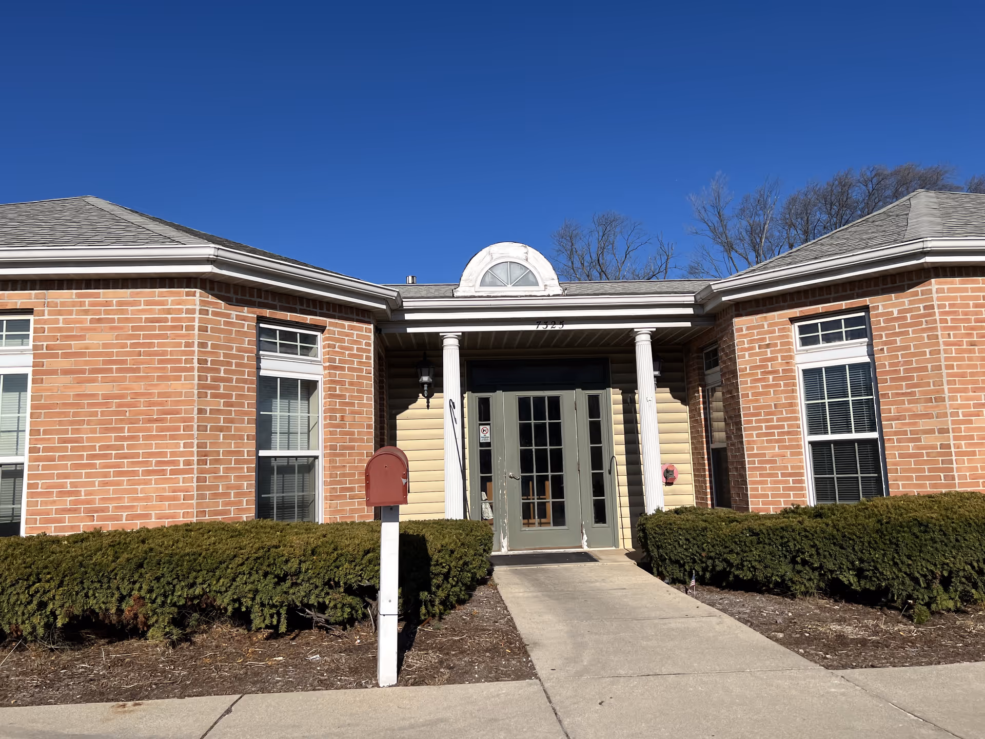 Front entrance of a single-story brick senior living building with double glass doors flanked by white columns, trimmed hedges, and a red mailbox.