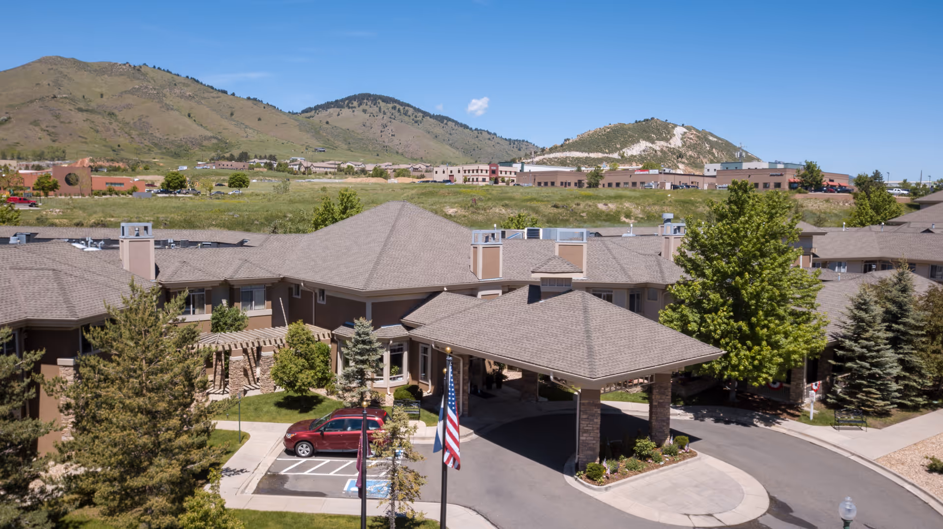 Aerial view of Golden Pond Senior Living facility showing a large building with a covered entrance, surrounded by trees and landscaping. In the background, there are hills and a clear blue sky.