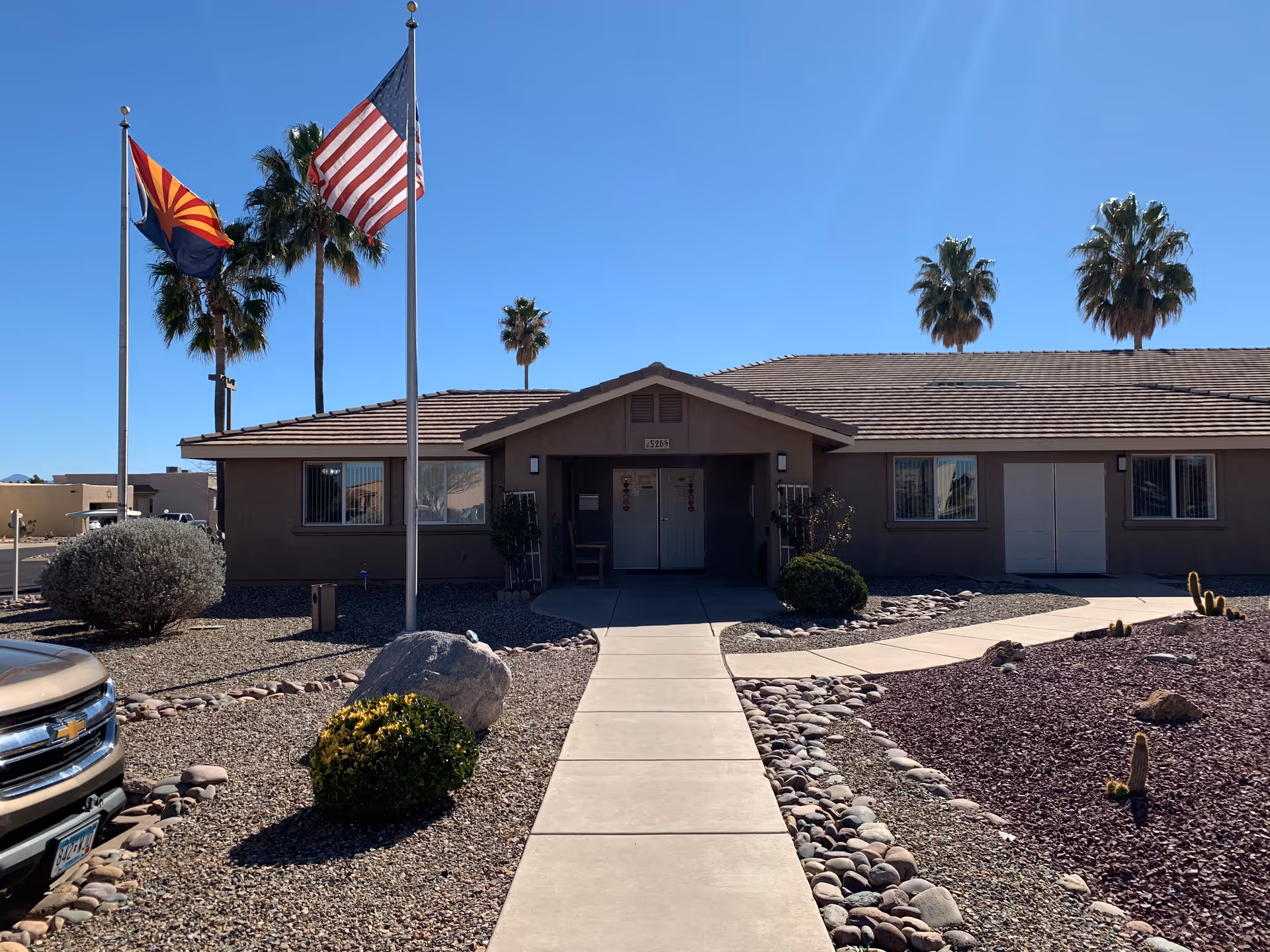 Single-story beige building entrance with American and Arizona flags, palm trees, and desert rock landscaping.