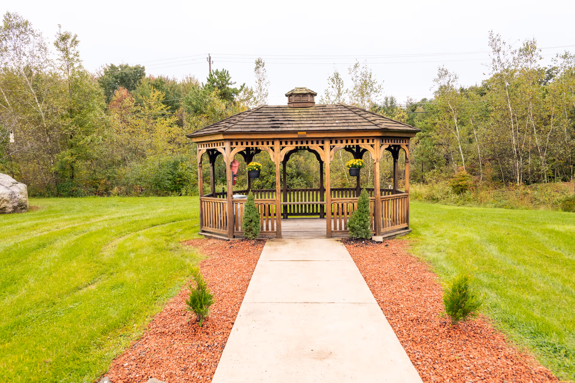 Wooden gazebo at the end of a concrete path surrounded by grass and trees.