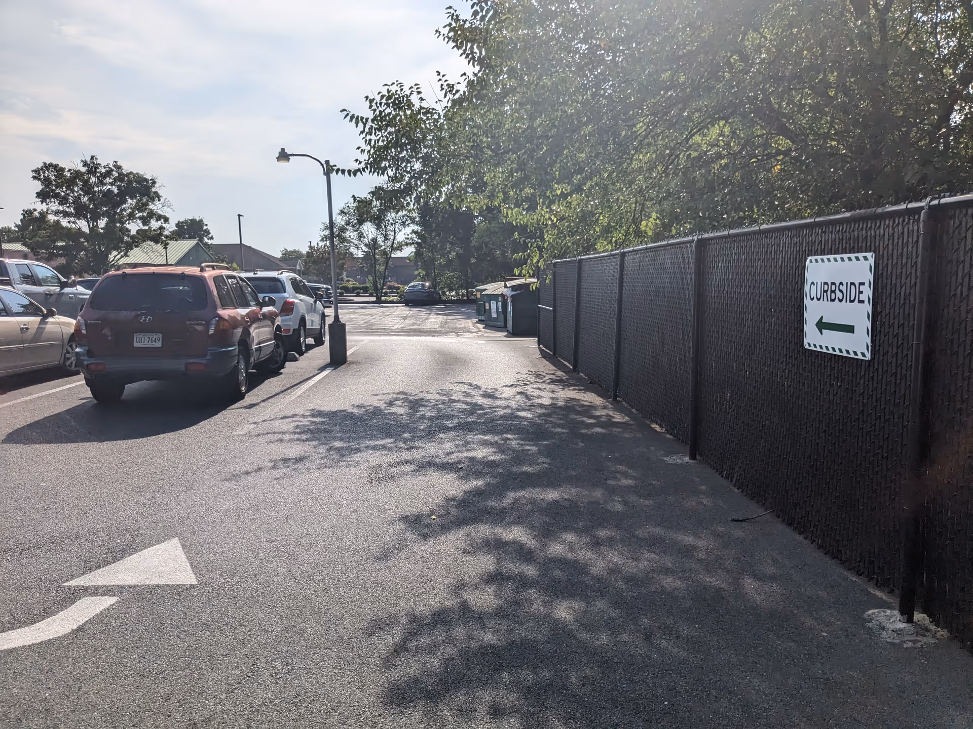 Parking lot beside a chain-link fence, with curbside pickup signage and trees.