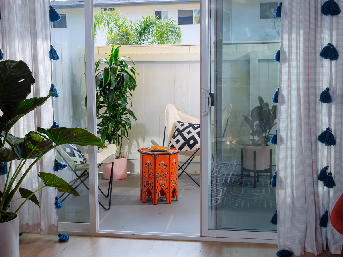 View through a sliding glass door to a small outdoor patio area with two beige butterfly chairs, one with a black and white geometric pillow, and a small red decorative table with a plate of food on it. There are potted plants on the patio and inside near the door, with white curtains featuring blue tassels framing the door.