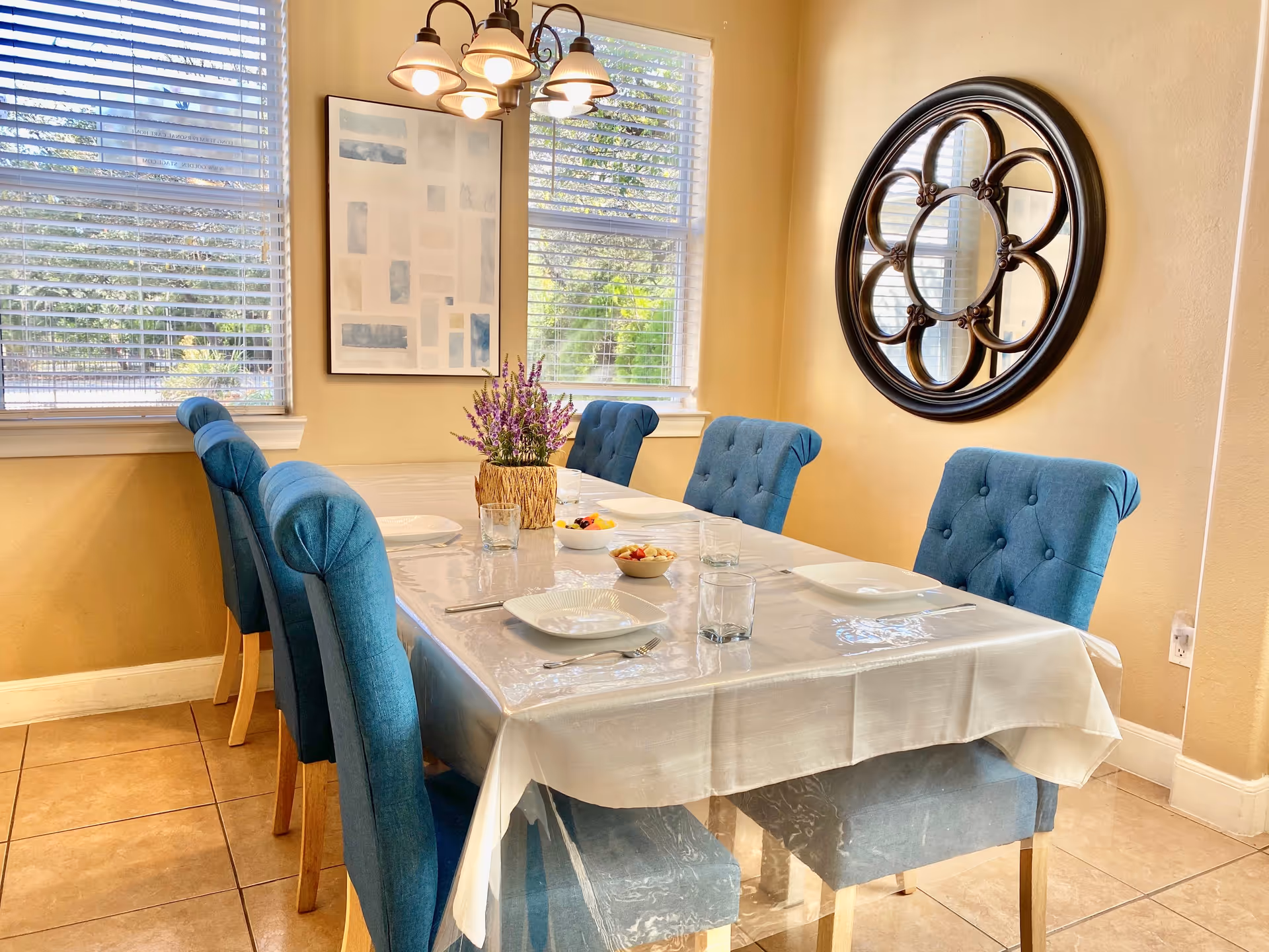 A dining room with a rectangular table covered with a white tablecloth and a clear protective cover. The table is set with white plates, glasses, and cutlery, and has a centerpiece of purple flowers in a woven basket. Six blue upholstered chairs with wooden legs surround the table. The room has beige walls, two large windows with white blinds, a framed abstract painting, and a decorative round mirror on the wall.