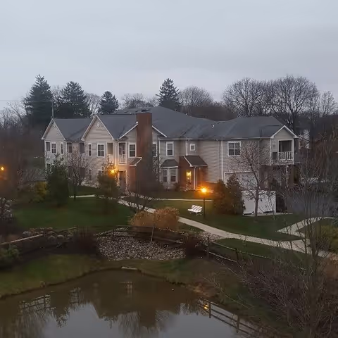 Two-story beige senior living building with balconies and lit exterior lamps beside a pond and walkways under an overcast sky.