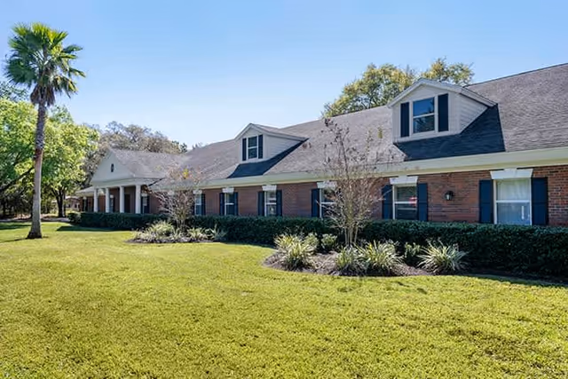 Exterior view of a single-story brick building with multiple windows and dormer windows on the roof, surrounded by a well-maintained lawn, shrubs, and a palm tree under a clear blue sky.