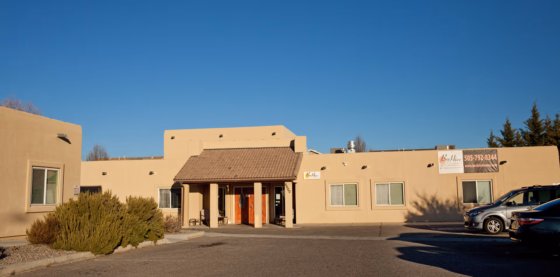 Exterior view of BeeHive Homes of Taylor Ranch, a single-story beige building with a tiled roof entrance, several windows, and a parking area with cars under a clear blue sky.