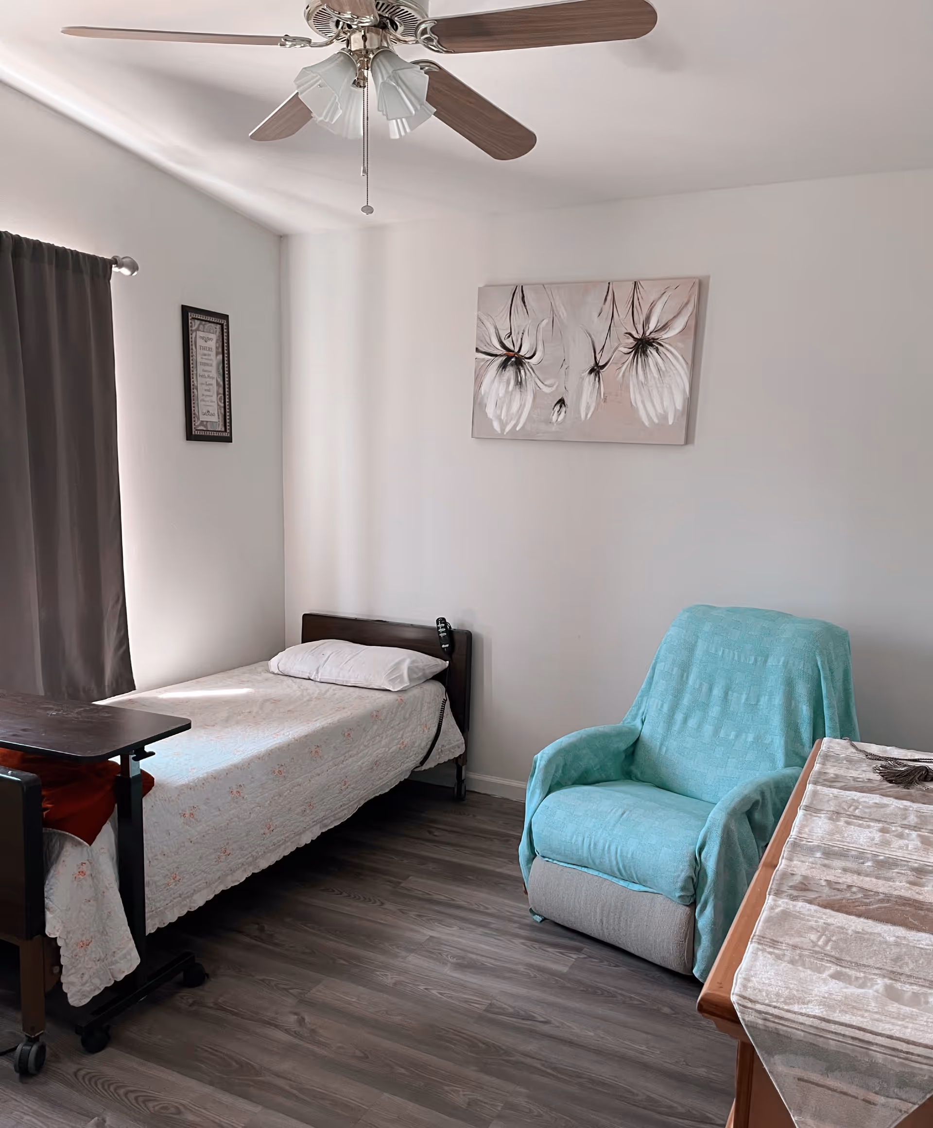 A simple bedroom with a single bed covered with a white floral bedspread, a pillow, and a remote control resting on the bed. Next to the bed is a dark wooden overbed table. A light blue armchair with a matching cover is positioned near a wooden dresser with a beige runner. The room has light-colored walls, a ceiling fan with wooden blades, a window with dark curtains, and two pieces of wall art featuring flowers and text.