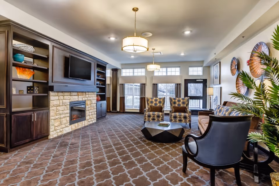 Spacious senior living lounge with patterned carpet, fireplace and TV, built-in shelving, and a seating area with chairs and a coffee table.